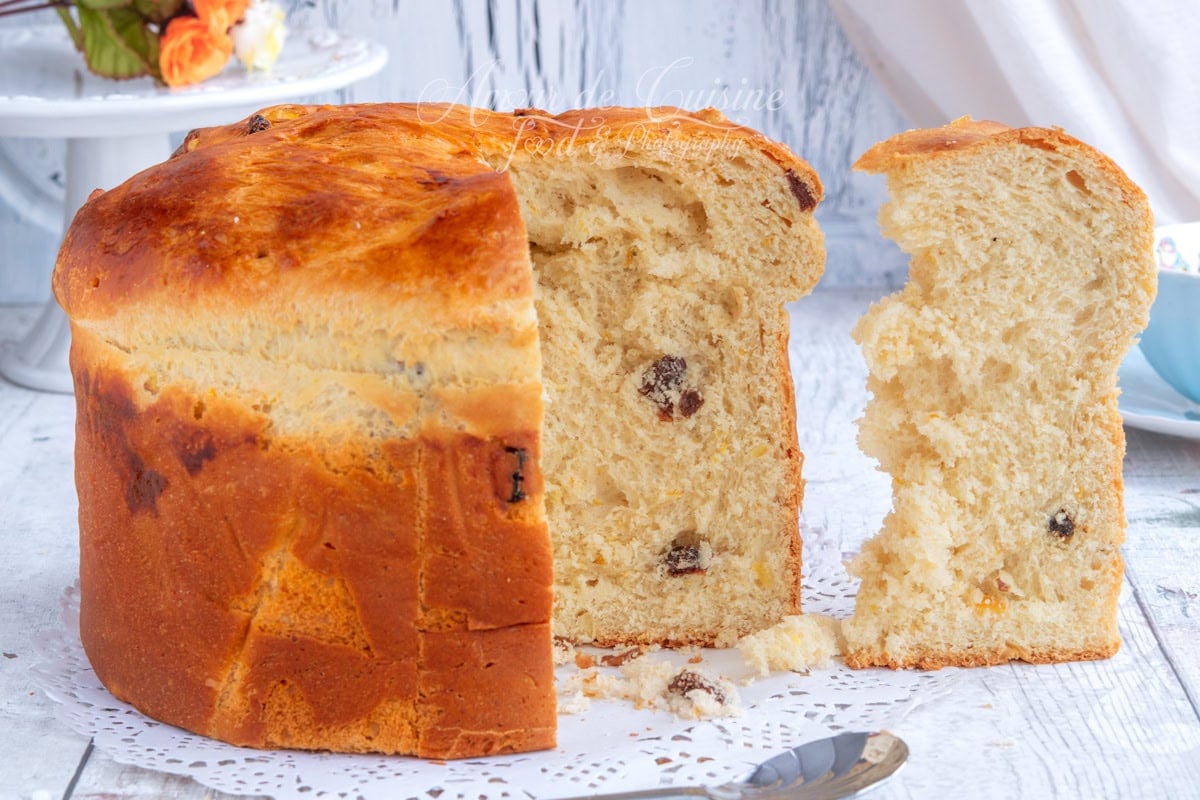 Panettone maison doré, coupé en deux avec une tranche servie à côté, montrant une mie moelleuse et quelques raisins secs, sur un napperon en papier.