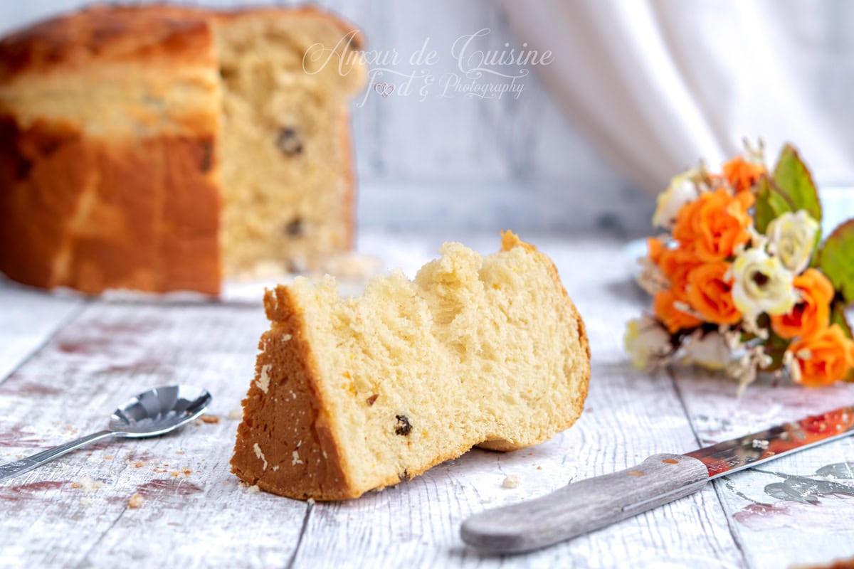 Part de panettone maison posée sur une table en bois, mie moelleuse avec quelques raisins, avec un couteau et une cuillère, et le panettone entier flou en arrière-plan.