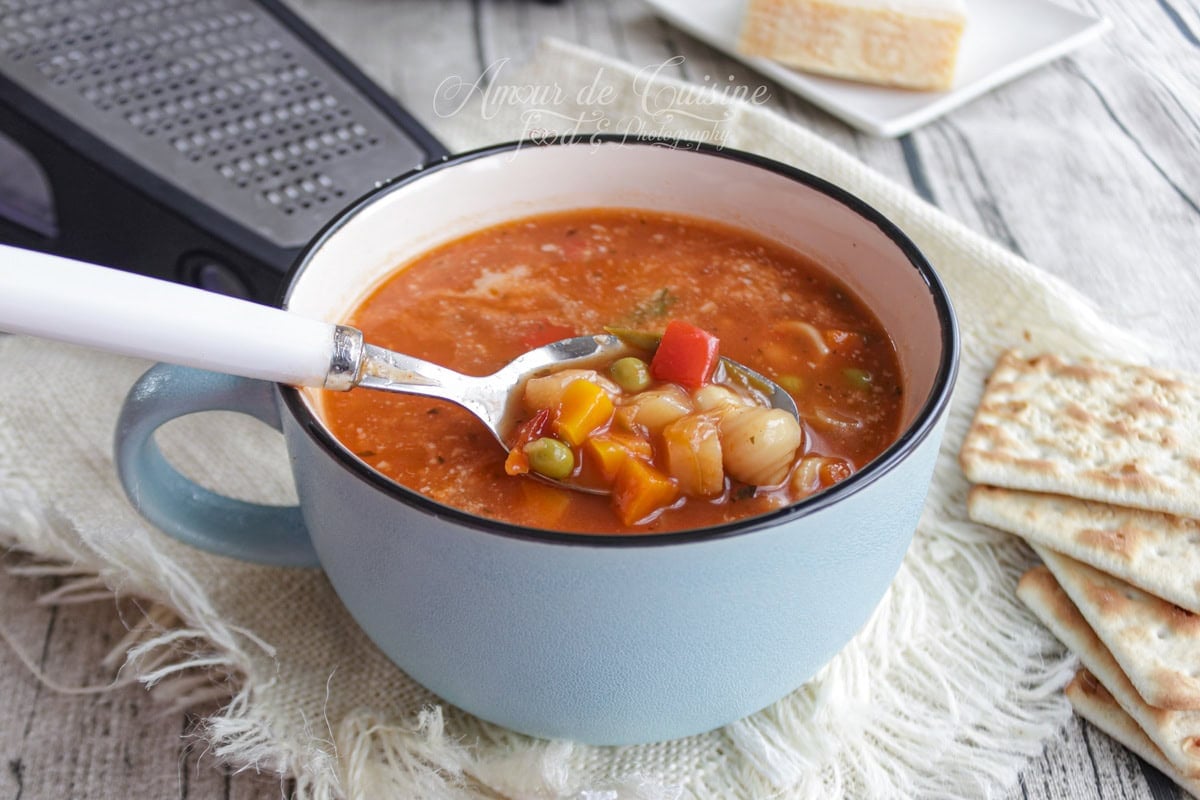 Soupe minestrone à la tomate servie dans une tasse bleue, avec une cuillère montrant des légumes en dés, petits pois et pâtes, accompagnée de crackers, sur un linge rustique.