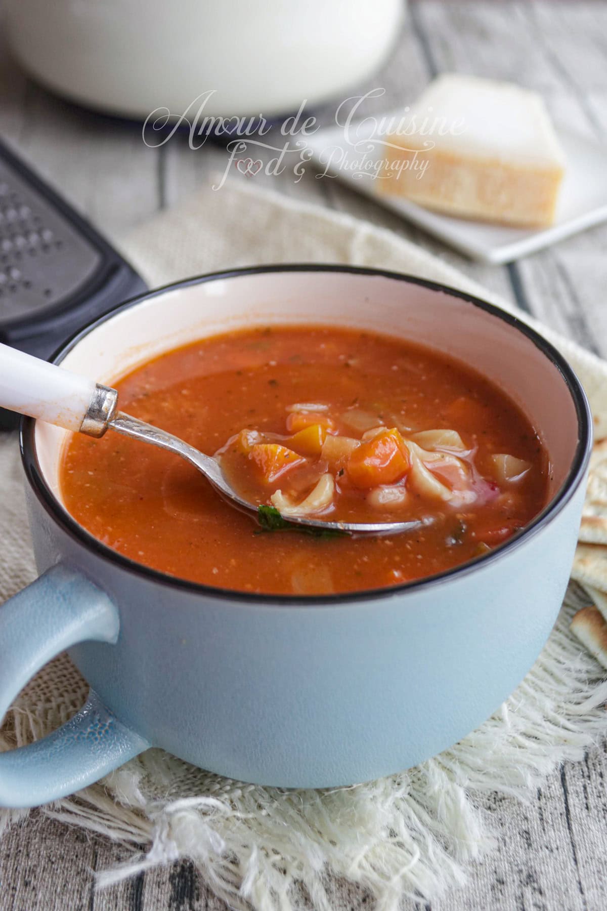 Bol de soupe minestrone à la tomate, avec légumes en dés et petites pâtes, servi dans une tasse bleue avec une cuillère, crackers et parmesan en arrière-plan.