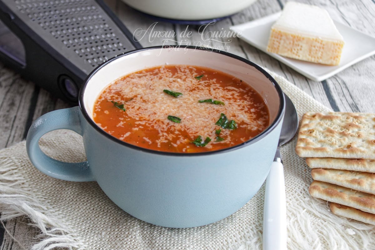 Tasse bleue de soupe minestrone à la tomate, parsemée de parmesan râpé et de basilic, posée sur un torchon, avec des crackers, une râpe et un morceau de fromage en arrière-plan.