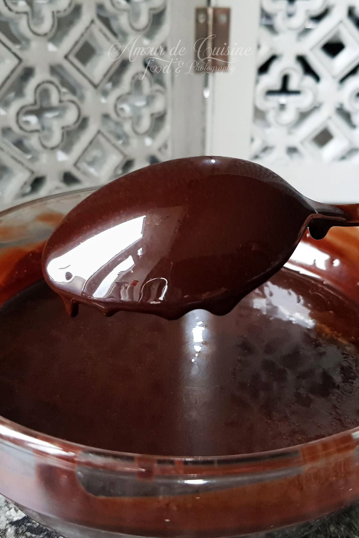 Ultra-glossy cocoa mirror glaze dripping from a spoon into a glass bowl, showing a smooth, reflective finish.