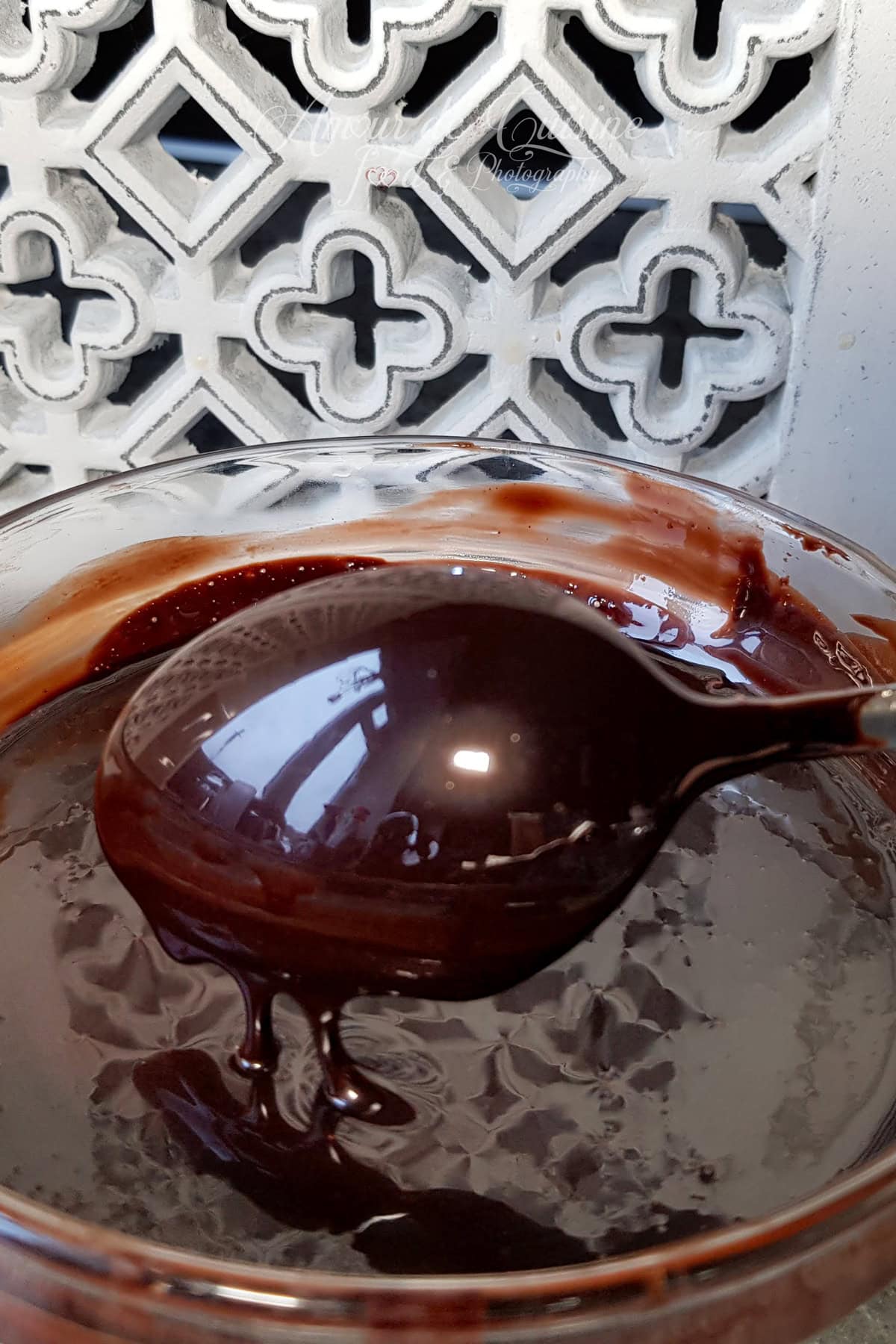 Spoon lifting ultra-glossy cocoa mirror glaze above a glass bowl, with a thick ribbon dripping back down and a reflective, mirror-like shine.