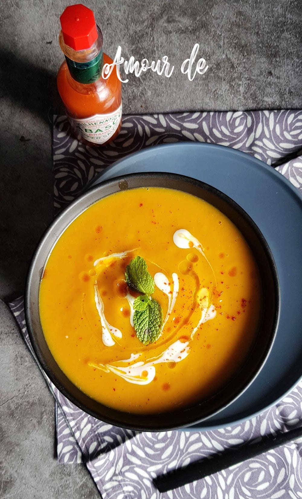 Bowl of creamy butternut squash soup, topped with fresh cream and mint leaves, placed on a blue plate with a patterned tablecloth, served alongside a bottle of hot sauce.