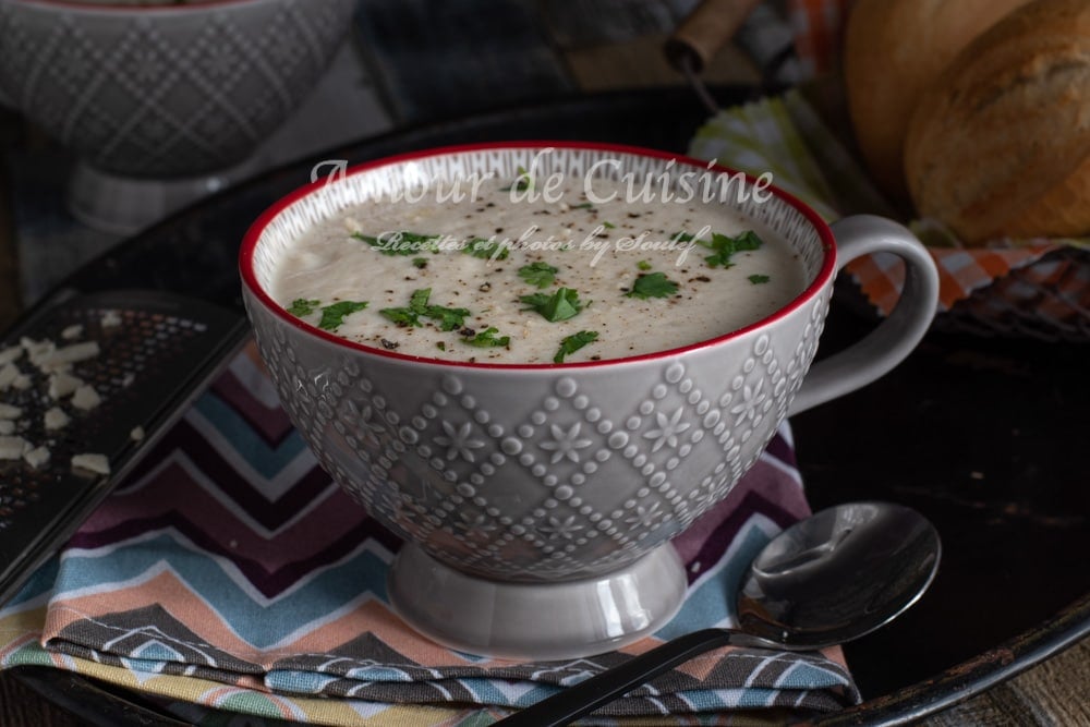 Bowl of creamy cauliflower soup topped with chopped parsley and cracked pepper, served in a patterned grey bowl with a red rim, placed on a colorful napkin beside a spoon and bread in the background.