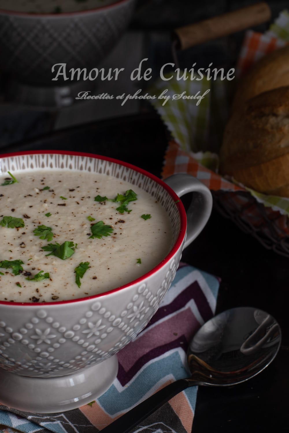 Close-up of creamy cauliflower soup in a patterned bowl with a red rim, sprinkled with black pepper and chopped parsley, set on a colorful napkin with a spoon and crusty bread in the background.