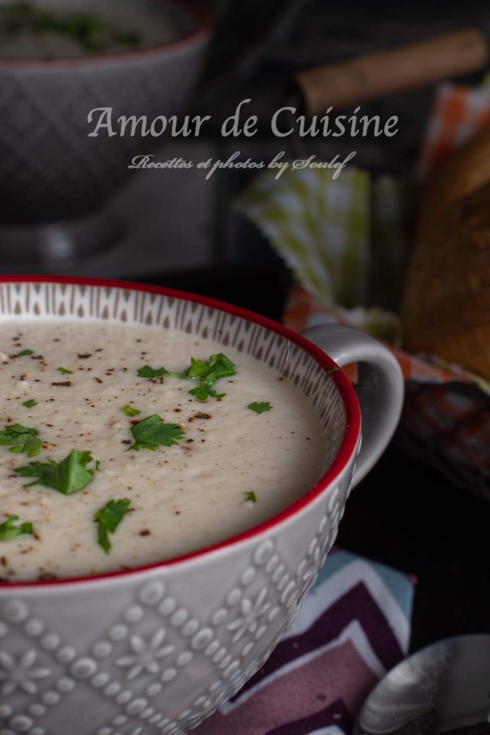 Close-up of creamy cauliflower soup in a grey patterned bowl with a red rim, topped with chopped parsley and black pepper, with crusty bread and a spoon softly blurred in the background.