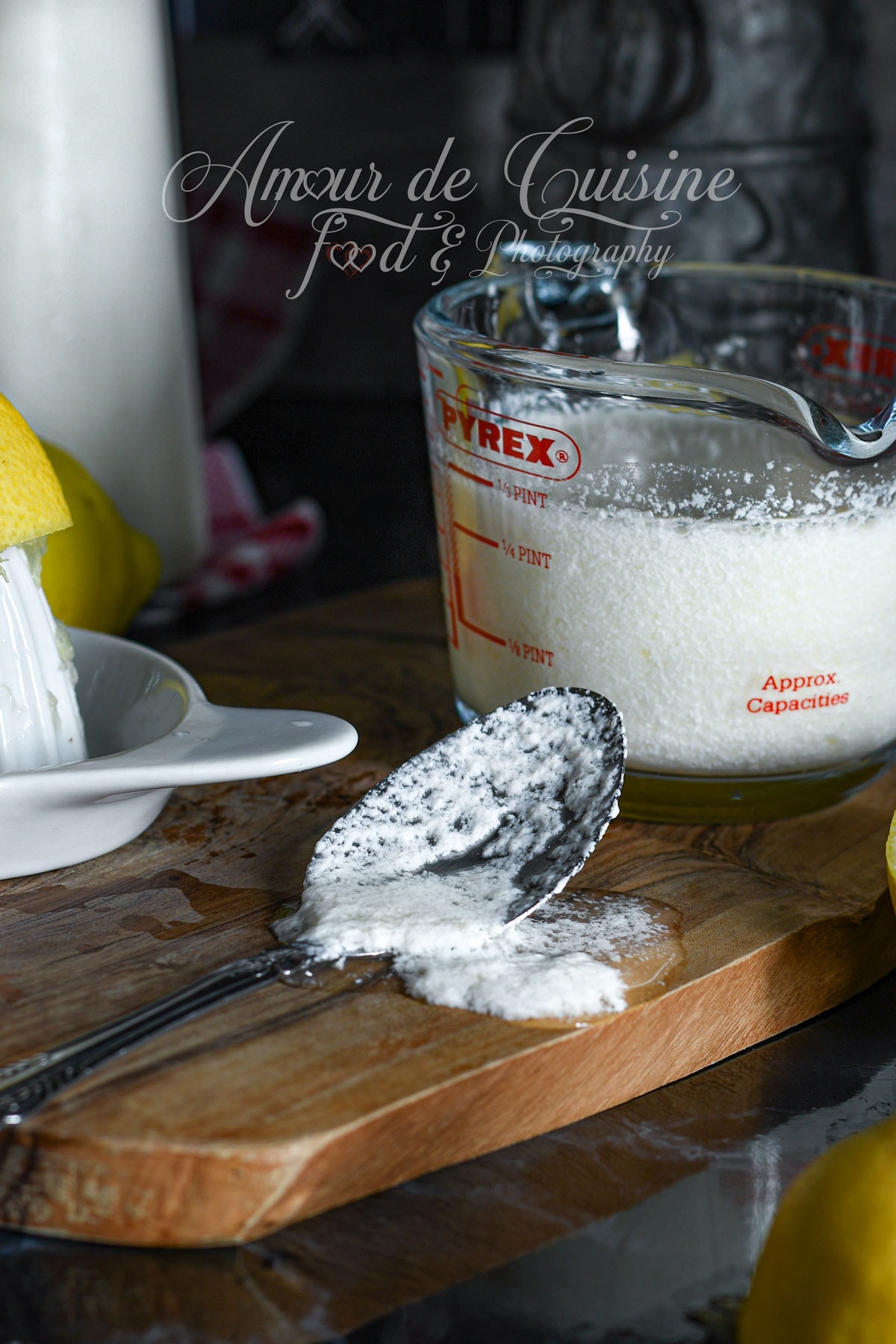 Spoonful of homemade buttermilk above a Pyrex measuring cup filled with fermented milk, freshly squeezed lemon and ceramic citrus juicer on a wooden board, cozy homemade kitchen vibe.