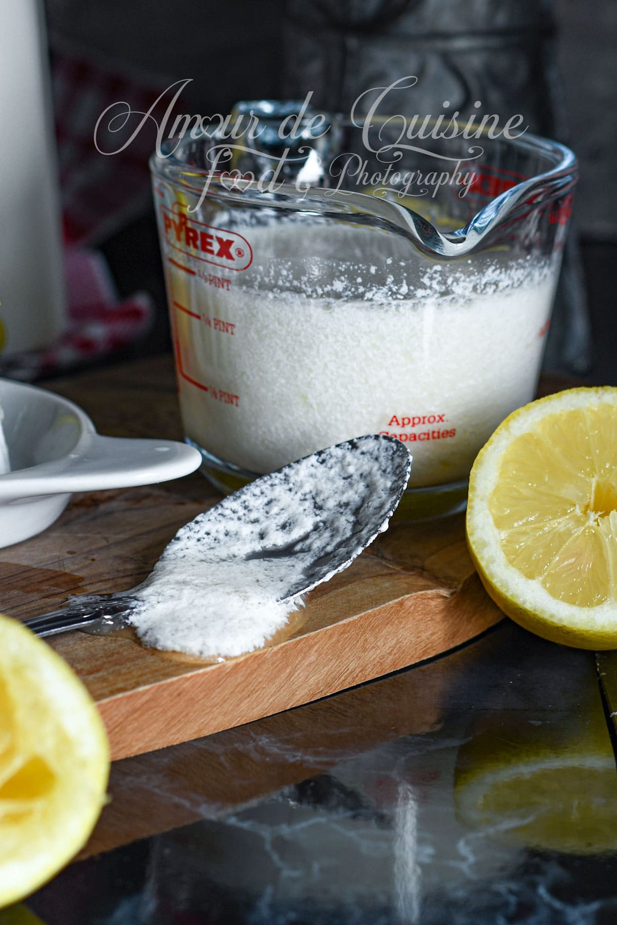Spoon covered with homemade buttermilk spilling onto a wooden board, next to a Pyrex measuring cup filled with fermented milk and a fresh lemon half, cozy kitchen preparation scene.