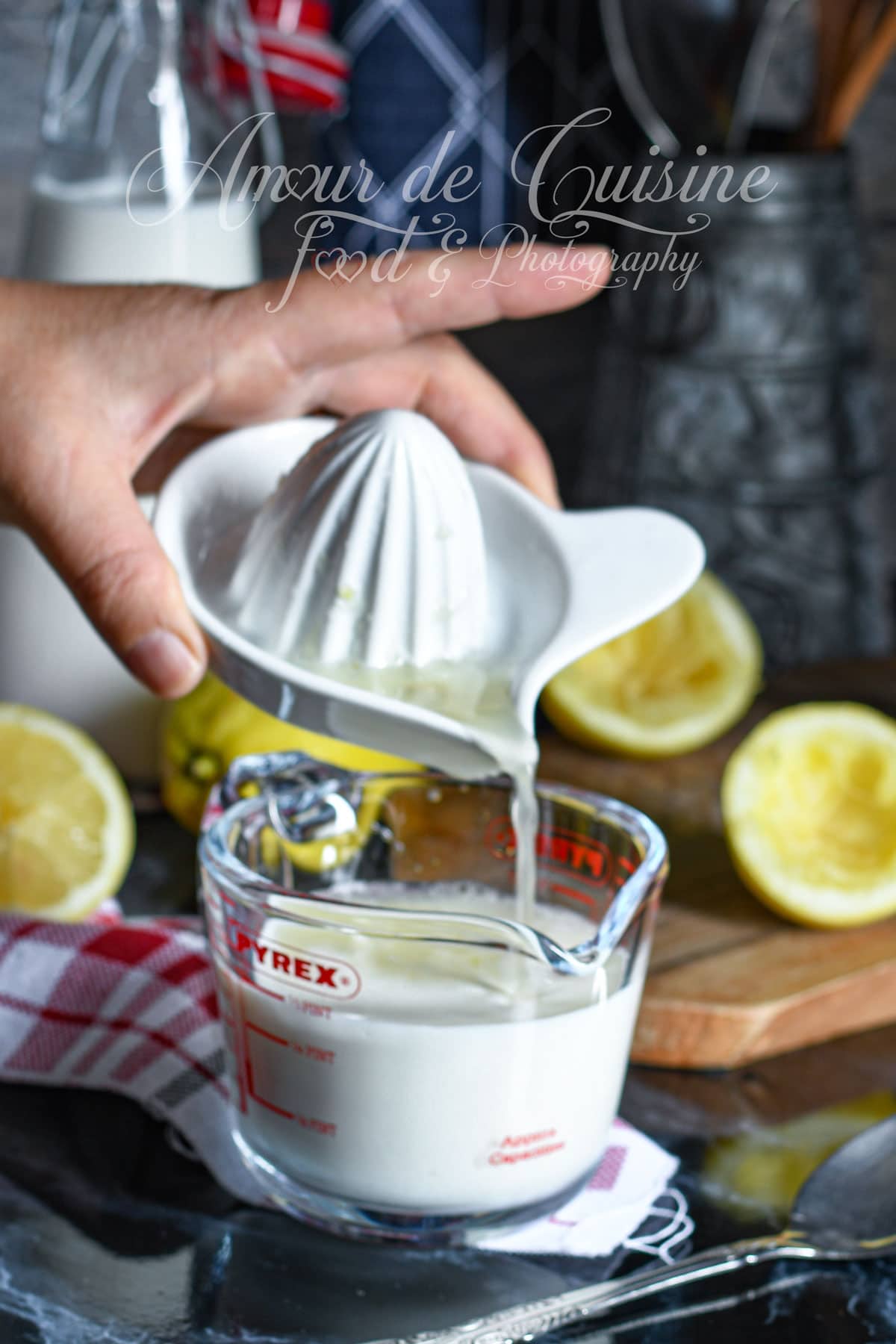 Hand squeezing a lemon with a ceramic juicer, pouring the juice into a Pyrex measuring cup filled with milk to make homemade buttermilk, with sliced lemons in the background.