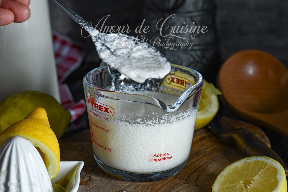 Cuillère de babeurre maison au-dessus d’un verre doseur Pyrex rempli de lait fermenté, citron fraîchement pressé et presse-agrumes en céramique sur une planche en bois, ambiance gourmande de cuisine maison.