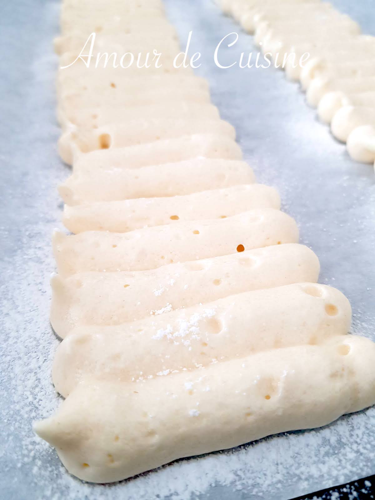 Ladyfinger batter piped into even strips on a parchment-lined baking tray, ready to be baked and generously dusted with powdered sugar.