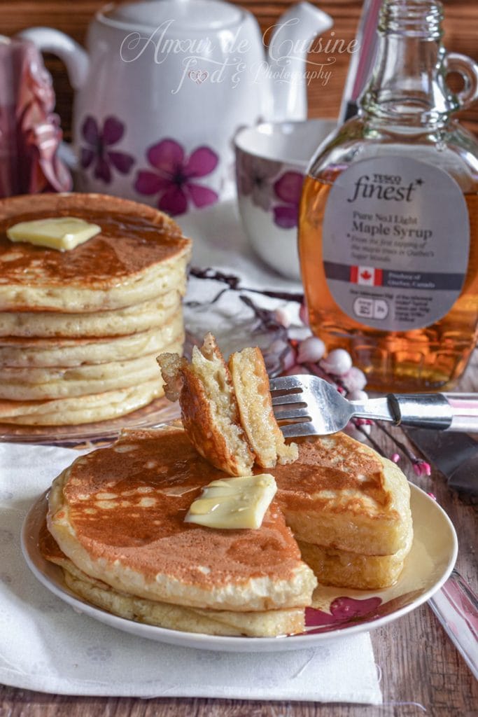 Plate of fluffy buttermilk pancakes topped with a melting pat of butter, with a cut piece held by a fork to show the texture, accompanied by a jar of maple syrup and a tea set in the background.
