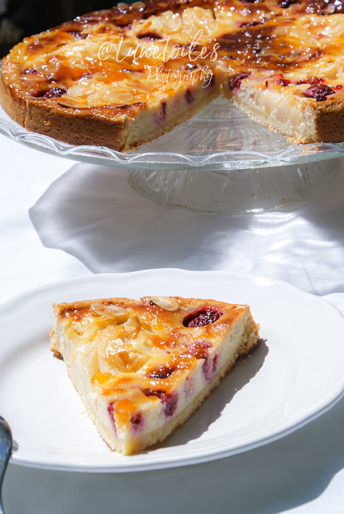 Tarte amandine aux poires et framboises sur un plat en verre, avec une part servie sur une assiette blanche, nappée de confiture et décorée d’amandes effilées
