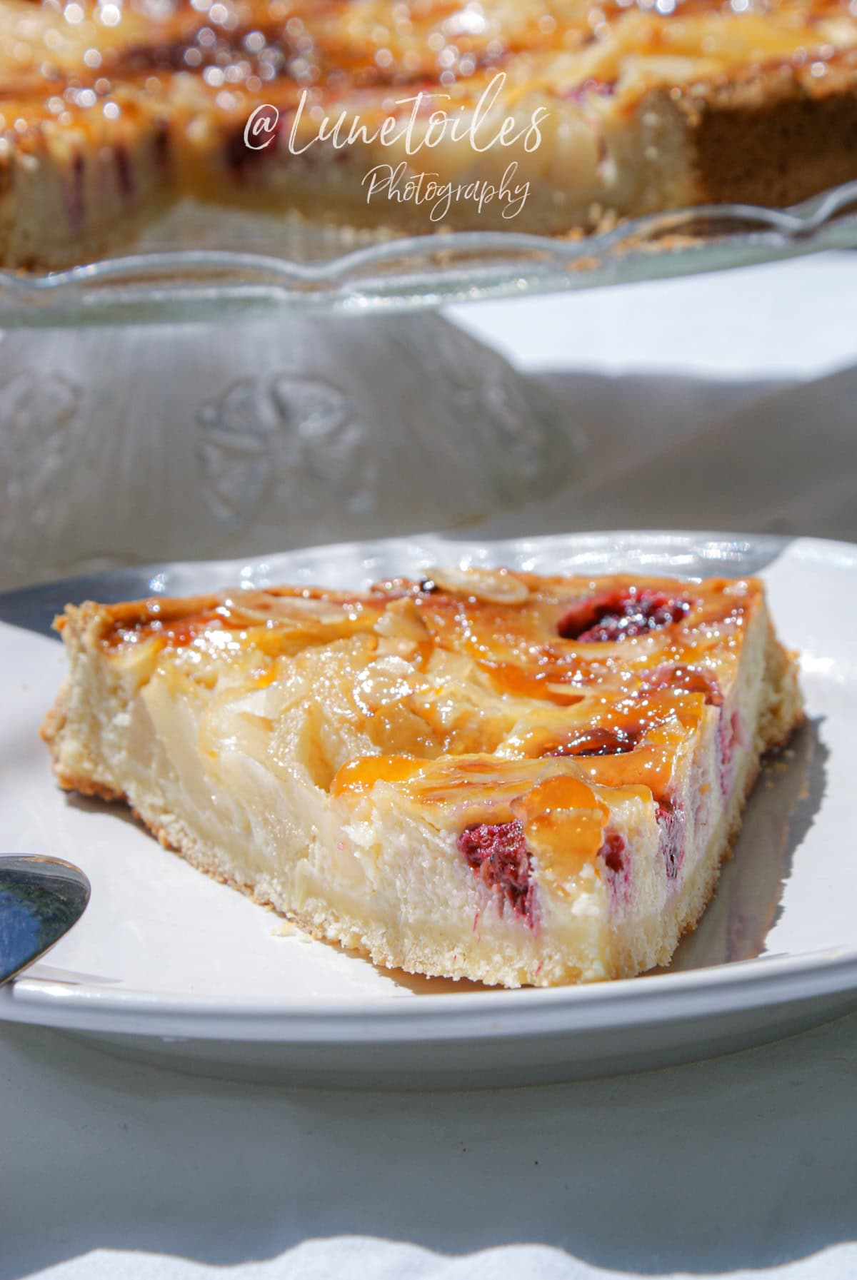 Part de tarte amandine aux poires et framboises nappée de confiture, servie sur une assiette blanche en plein soleil