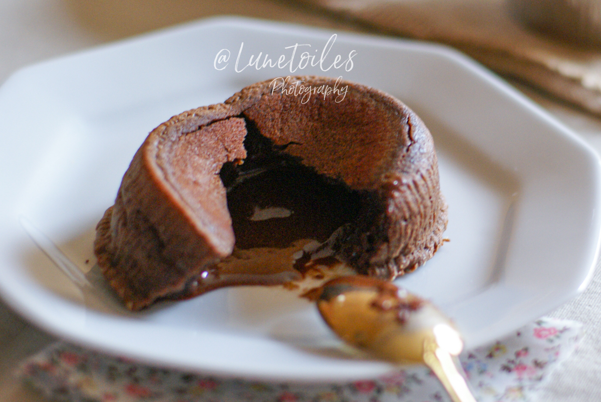 Moelleux au chocolat ouvert sur une assiette blanche, laissant couler son cœur fondant, avec une cuillère dorée posée devant et un tissu à motif floral en dessous de l’assiette