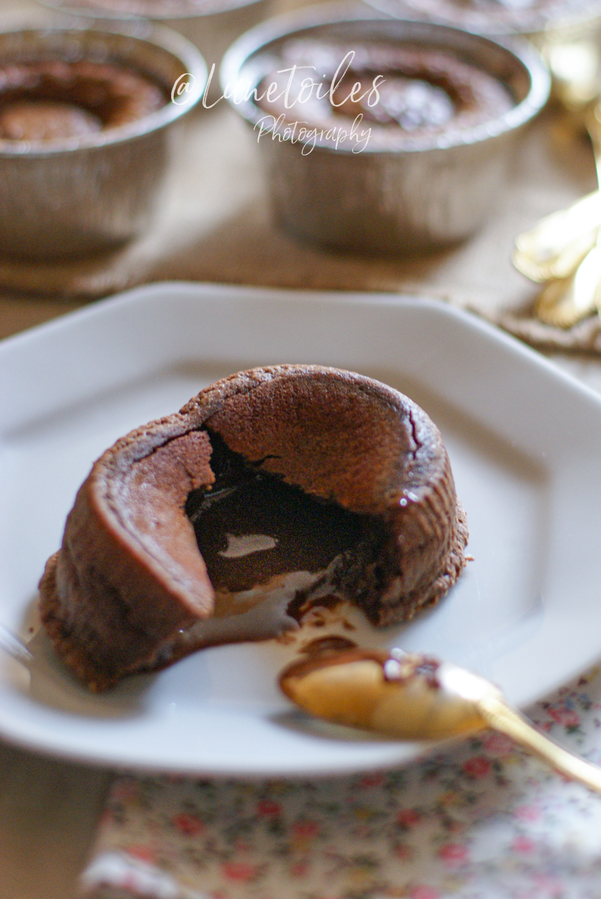 Moelleux au chocolat individuel ouvert sur une assiette blanche, laissant apparaître un cœur fondant coulant, avec une cuillère dorée posée à côté. En arrière-plan, plusieurs moules en aluminium contenant d’autres fondants cuits.
