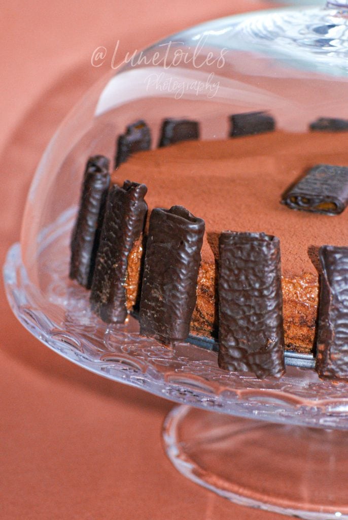 Flourless, gluten-free chocolate cake viewed from the side under a glass dome, decorated with chocolate-coated rolled biscuits around the edge, displayed on a clear cake stand.