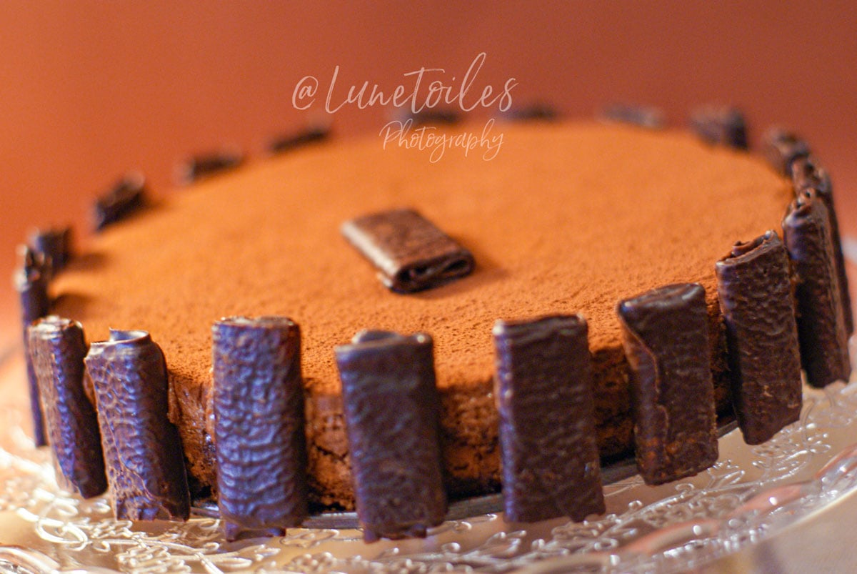 Gâteau au chocolat sans farine et sans gluten, décoré de rouleaux de chocolat tout autour et d’un carré de chocolat au centre, présenté sur un plat en verre.