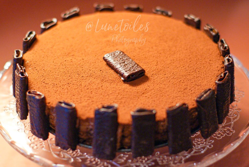 Flourless, gluten-free chocolate cake topped with cocoa powder and surrounded by chocolate-coated rolled biscuits, displayed on a decorative glass cake stand.