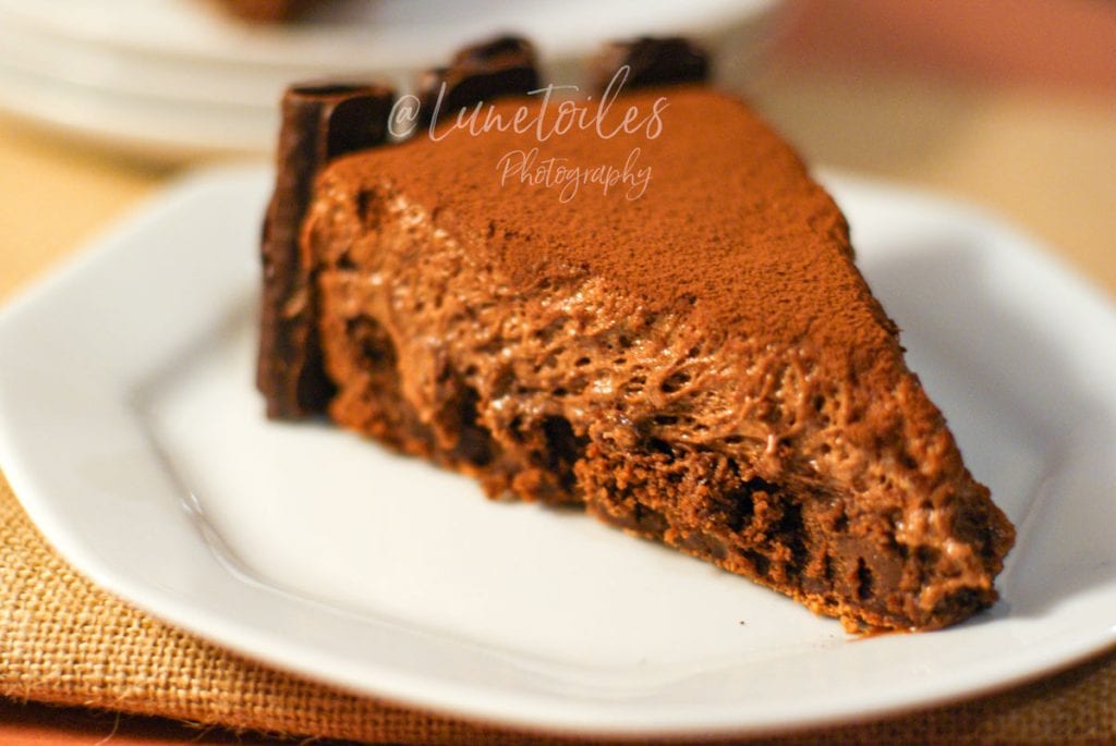 Slice of flourless, gluten-free chocolate cake with a rich, airy mousse texture, topped with cocoa powder and chocolate curls, served on a white plate.