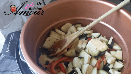 Légumes coupés – aubergines et poivrons rouges – en début de cuisson dans la cuve d’un Cookeo en cuivre, mélangés avec une cuillère en bois pour préparer une ratatouille maison.