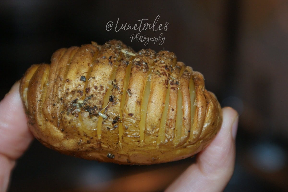 Roasted hasselback potato, golden and herb-seasoned, held between fingers with its thin slices fanned open.