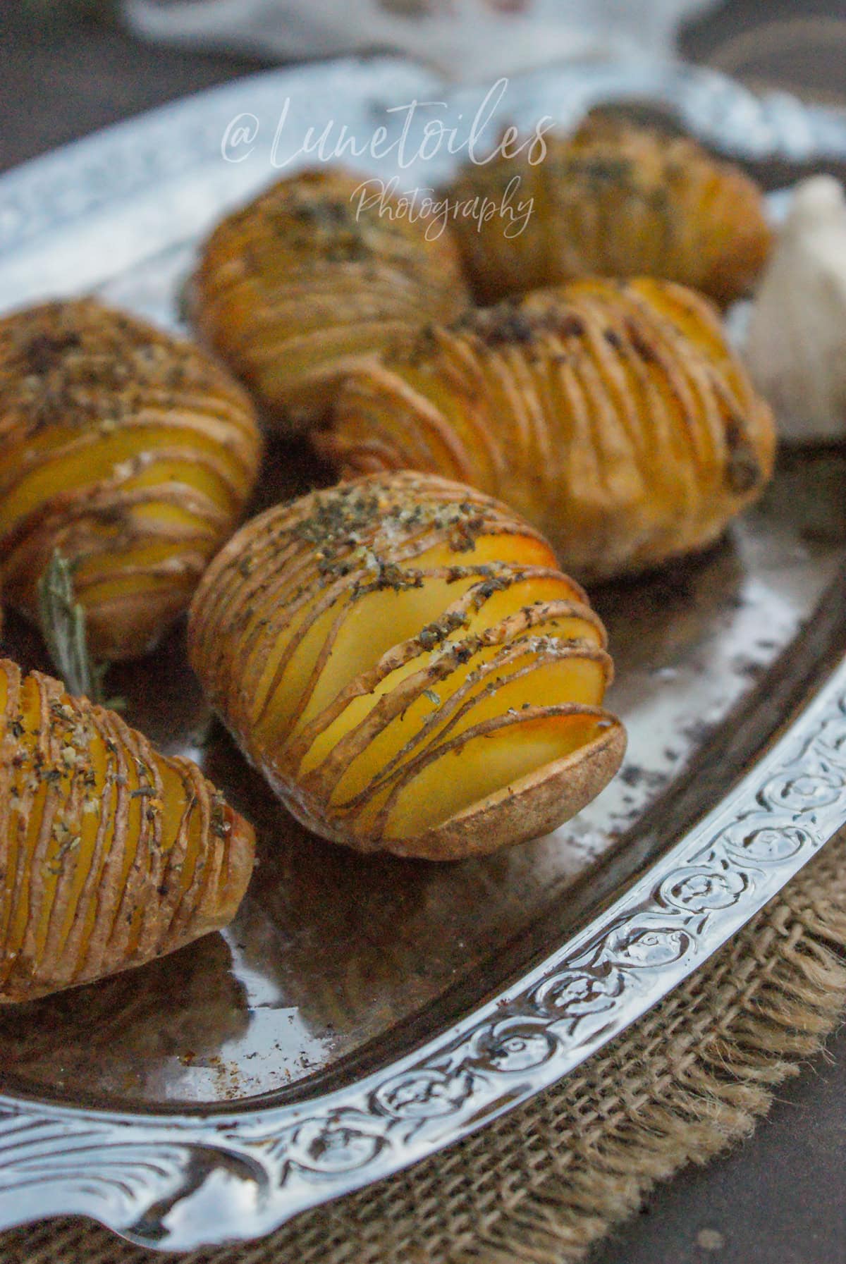 Oven-roasted hasselback potatoes sliced into thin fan-like layers, seasoned with herbs and olive oil, served on a silver platter.