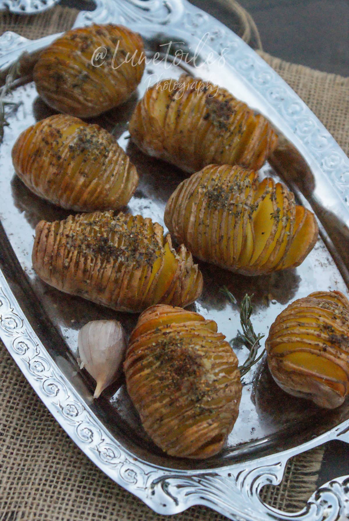 Golden roasted hasselback potatoes seasoned with herbs, arranged on a silver tray with a garlic clove and a sprig of rosemary.