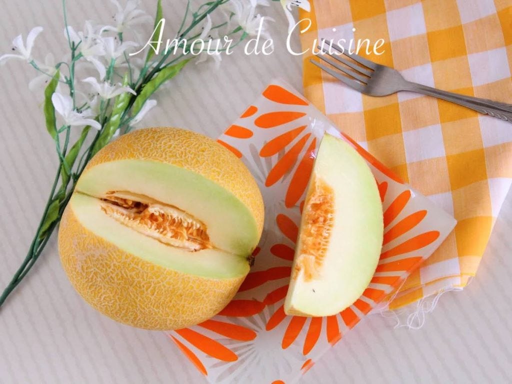 Fresh melon cut in half with visible seeds, placed next to a melon wedge on a floral-patterned plate, beside a yellow gingham napkin and fork, styled with white flowers