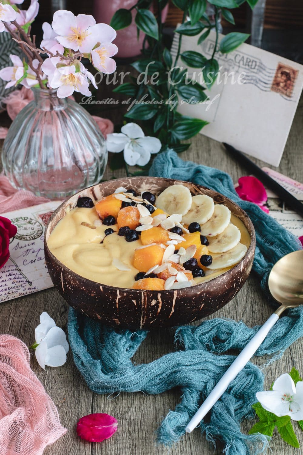 Mango banana smoothie bowl in a coconut bowl, topped with banana slices, mango chunks, blueberries, and flaked almonds, on a rustic table with a gold spoon, flowers, and pastel decor in the background.