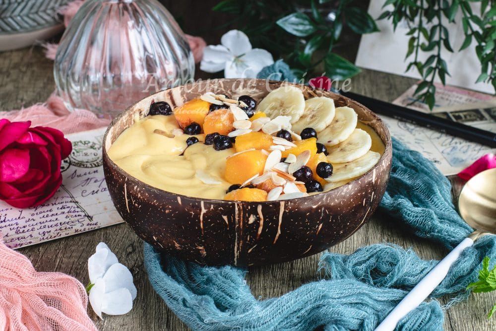 Close-up of a creamy mango banana smoothie bowl in a coconut bowl, topped with banana slices, mango chunks, blueberries, and flaked almonds, on a rustic table with a gold spoon.
