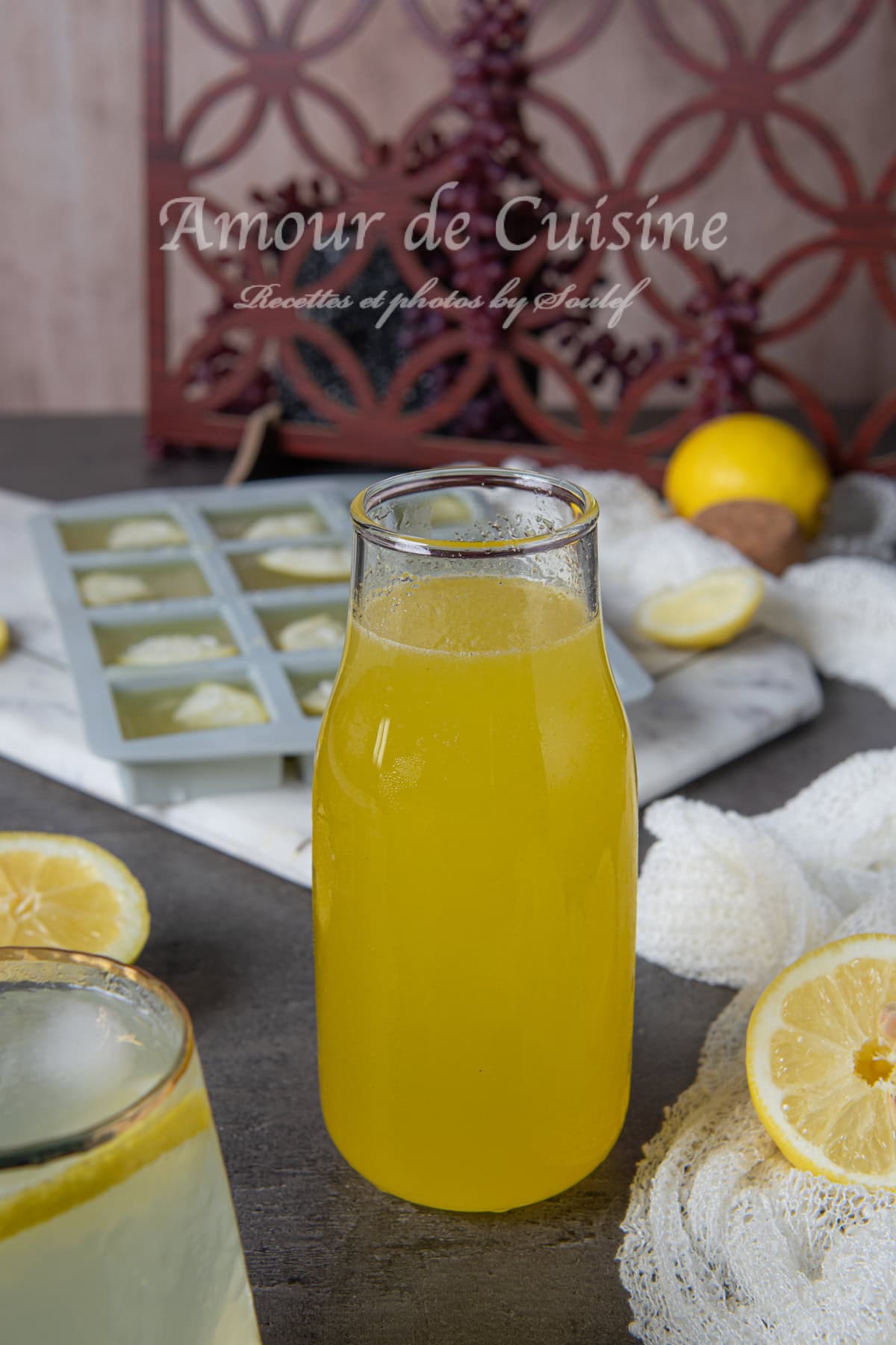 Bouteille en verre remplie de concentré de citron maison jaune, posée sur une table avec des citrons coupés, un bac à glaçons au fond et un verre de citronnade partiellement visible au premier plan.