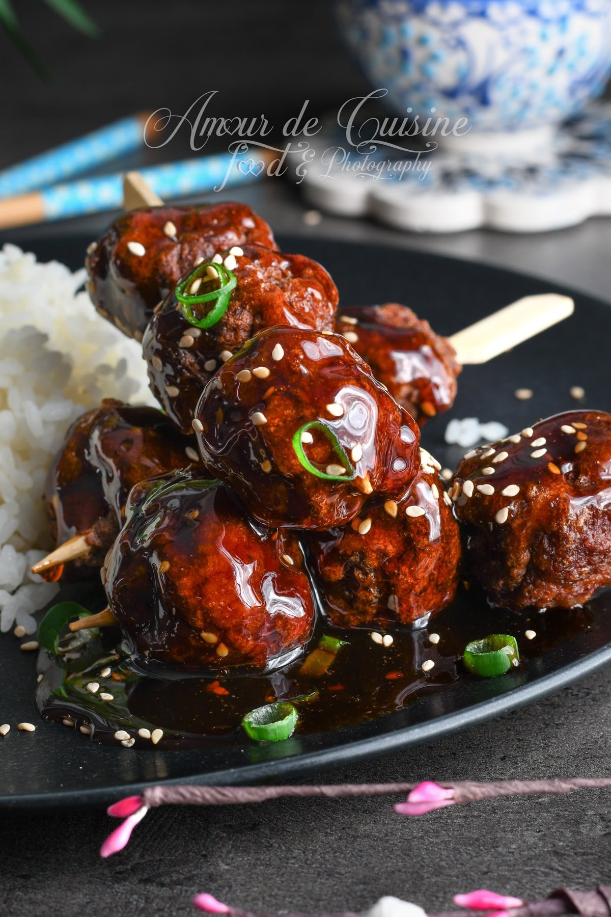 Brochettes de boulettes de poulet yakitori laquées de sauce teriyaki brillante, parsemées de sésame et d’oignon vert, servies avec du riz blanc sur une assiette noire.