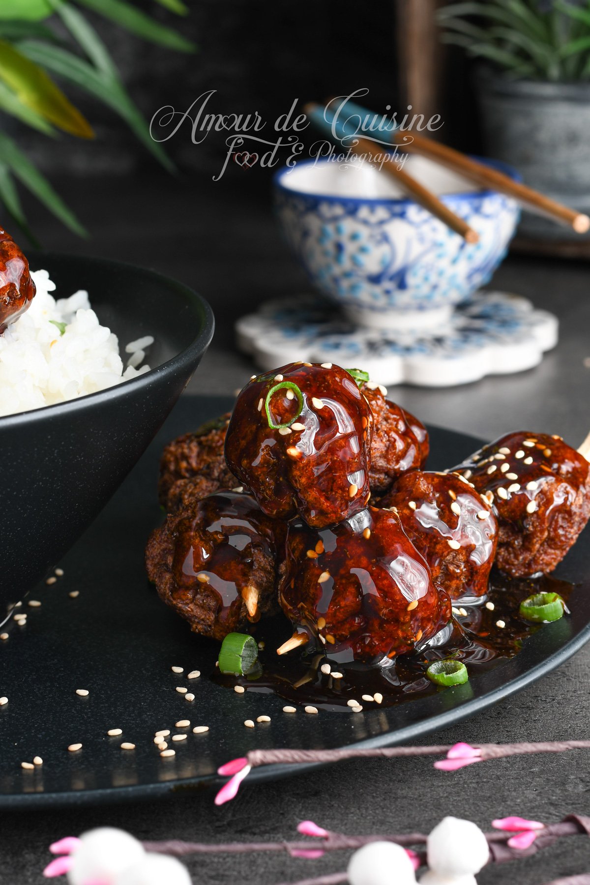 Brochettes de boulettes de poulet yakitori nappées de sauce teriyaki brillante, avec sésame et oignon vert, servies sur assiette noire à côté d’un bol de riz blanc.