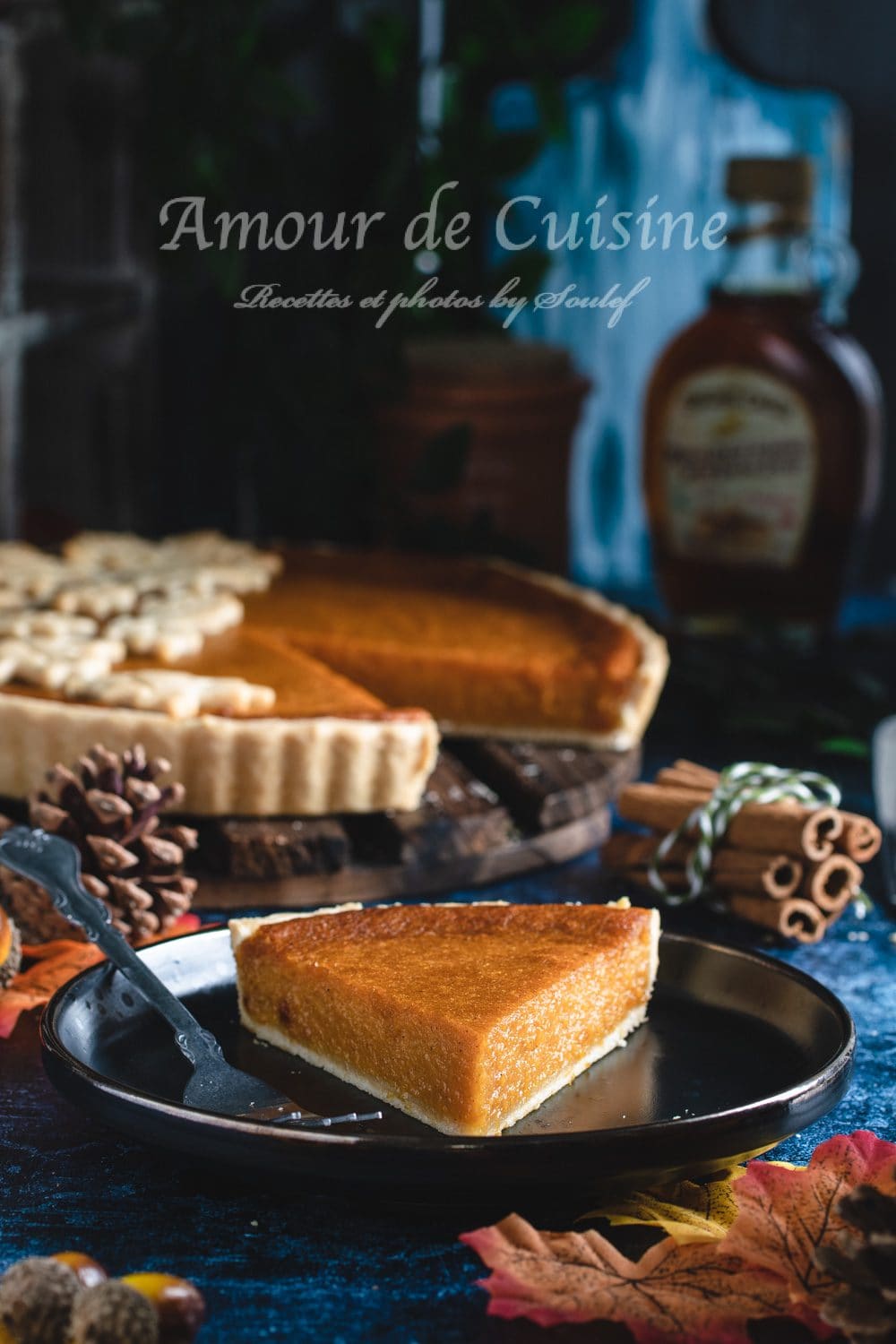 Slice of sweet potato maple pie served on a black plate, placed in front of the whole pie decorated with pastry leaves, set against an autumn backdrop of cinnamon sticks, pinecones, and maple syrup in the background