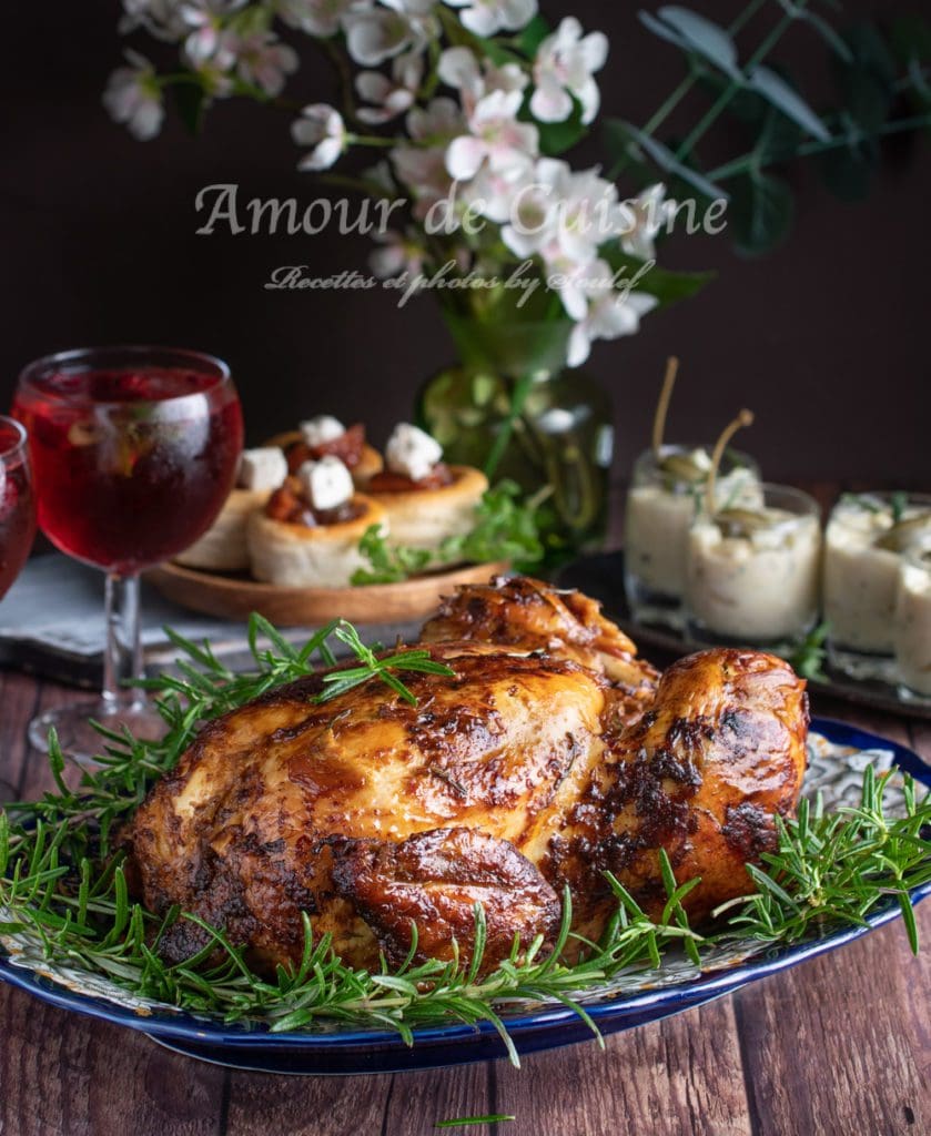 Golden oven-roasted capon on a serving platter, garnished with fresh rosemary, on a rustic table with a red drink and festive appetisers in the background.