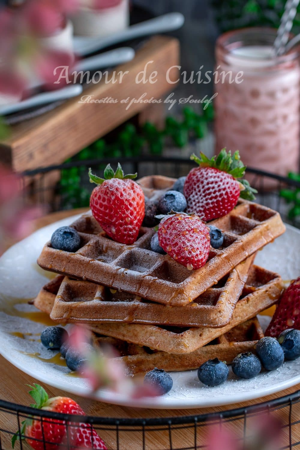 Stack of buckwheat waffles dusted with powdered sugar, topped with fresh strawberries and blueberries, with a drizzle of syrup on a white plate and pink drinks blurred in the background.