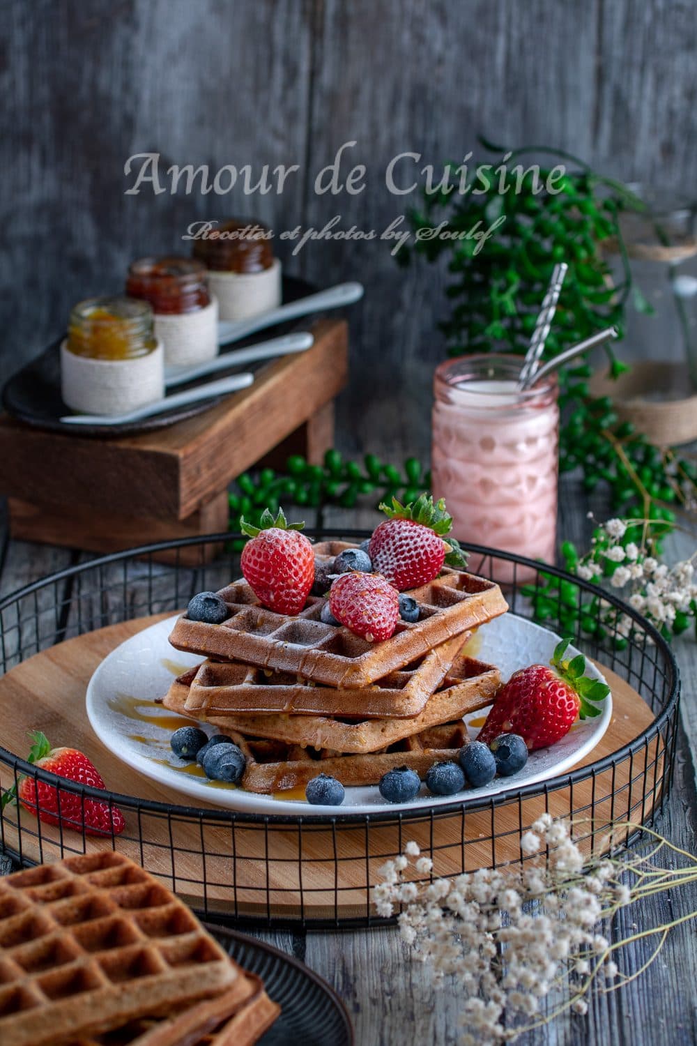 Buckwheat waffles stacked on a white plate with powdered sugar, strawberries and blueberries, drizzled with syrup, served on a wooden tray with jam jars and a pink drink in the background.