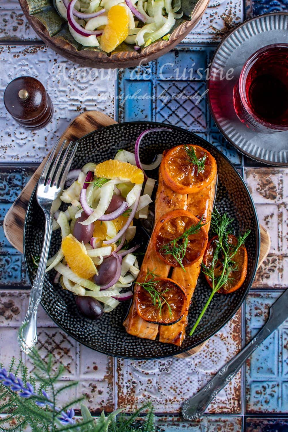 Oven-baked clementine-glazed salmon fillet, topped with caramelized slices and fresh dill, served with a crisp salad of fennel, oranges, olives, and red onions on a black plate.
