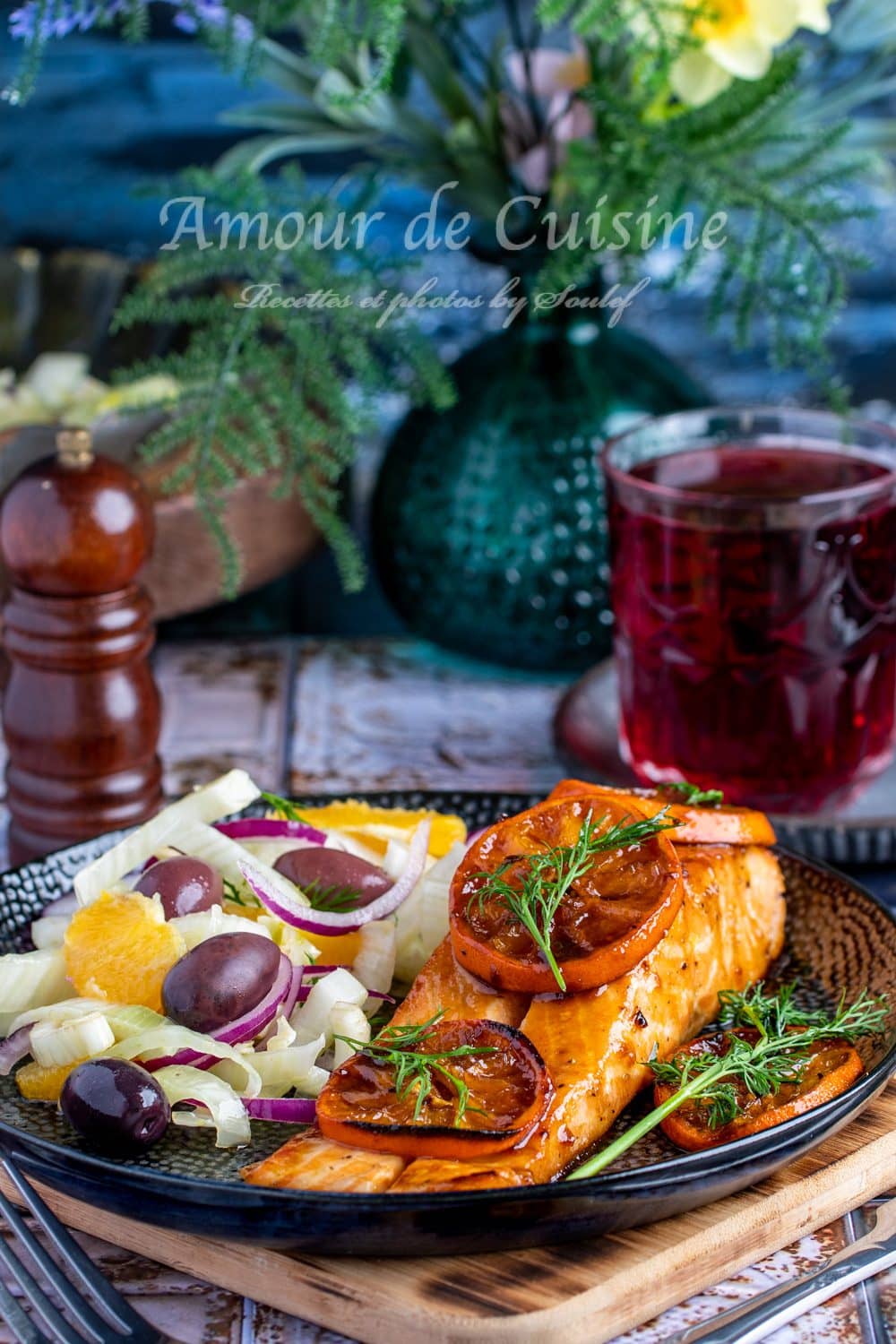 Pavé de saumon glacé à la clémentine, garni de tranches d’agrumes caramélisées et d’aneth frais, servi avec une salade de fenouil, oranges, olives et oignons rouges, sur une assiette noire élégante
