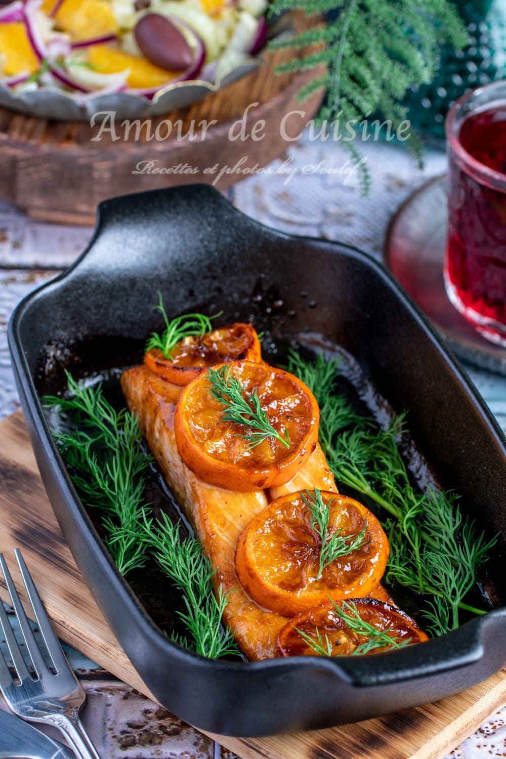 Oven-baked clementine-glazed salmon fillet, topped with caramelized citrus slices and fresh dill sprigs, presented in a black cast-iron dish on a wooden board.