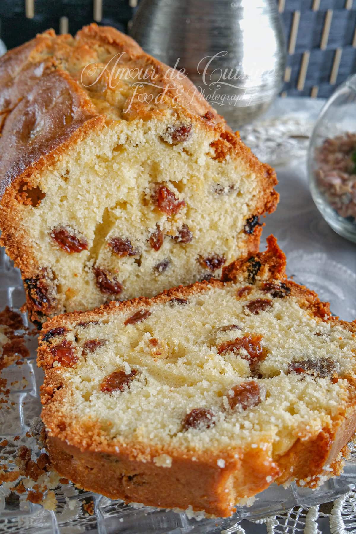 Close-up of a moist raisin loaf cake cut in half, with one slice in the foreground on a glass plate, crumbs scattered around and table décor in the background.