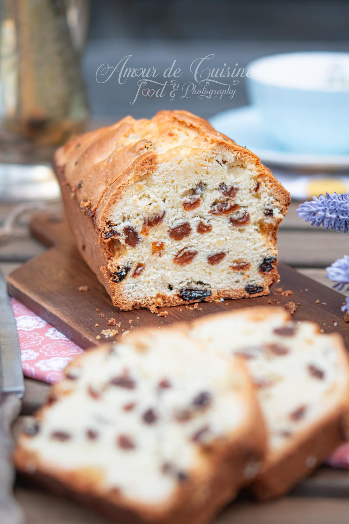 Moist raisin loaf cake sliced on a wooden board, with a blurred slice in the foreground and a teacup in the background.