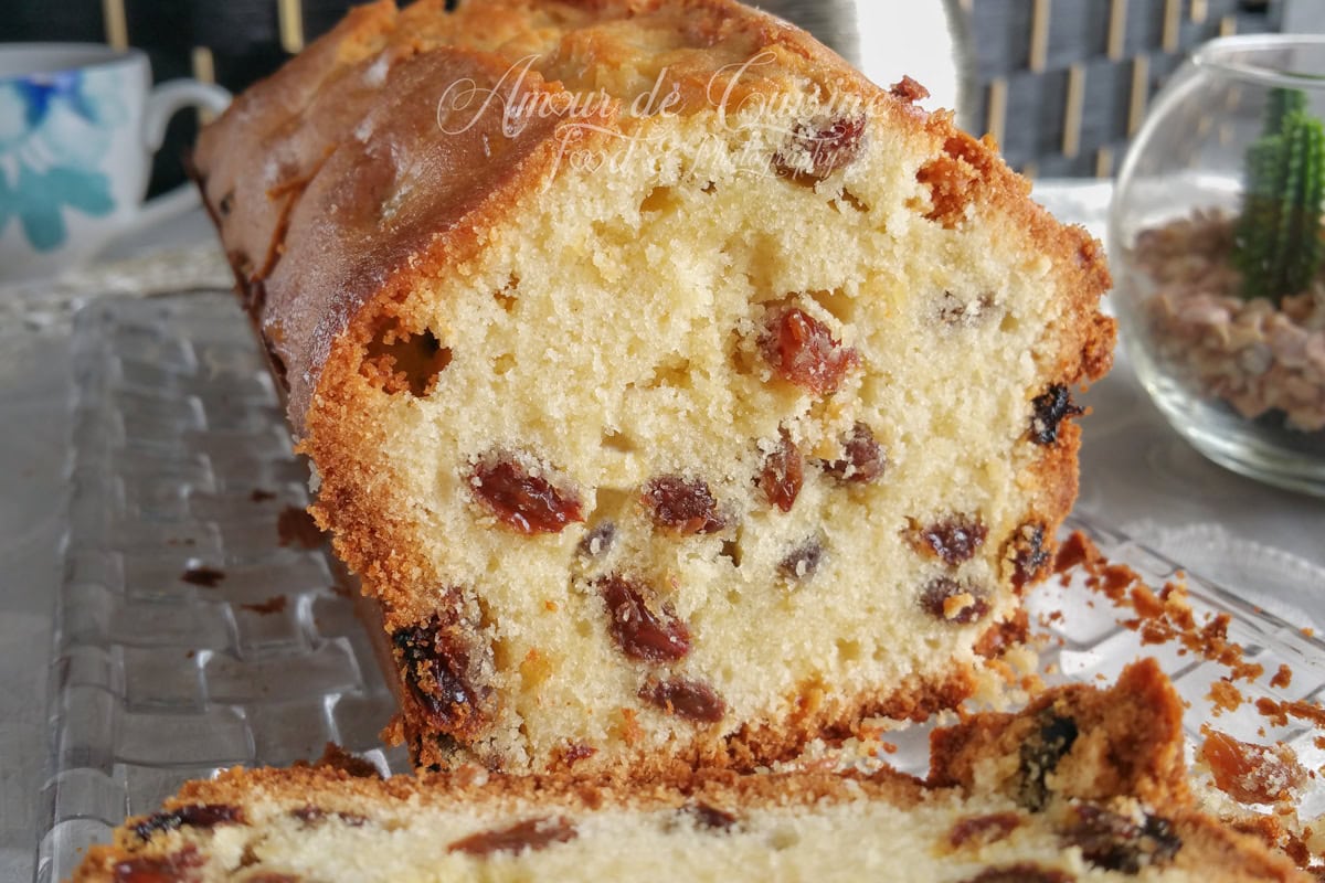 Close-up of a golden raisin loaf cake, sliced to show a moist crumb packed with raisins, with one slice in the foreground and a few crumbs on a glass platter.