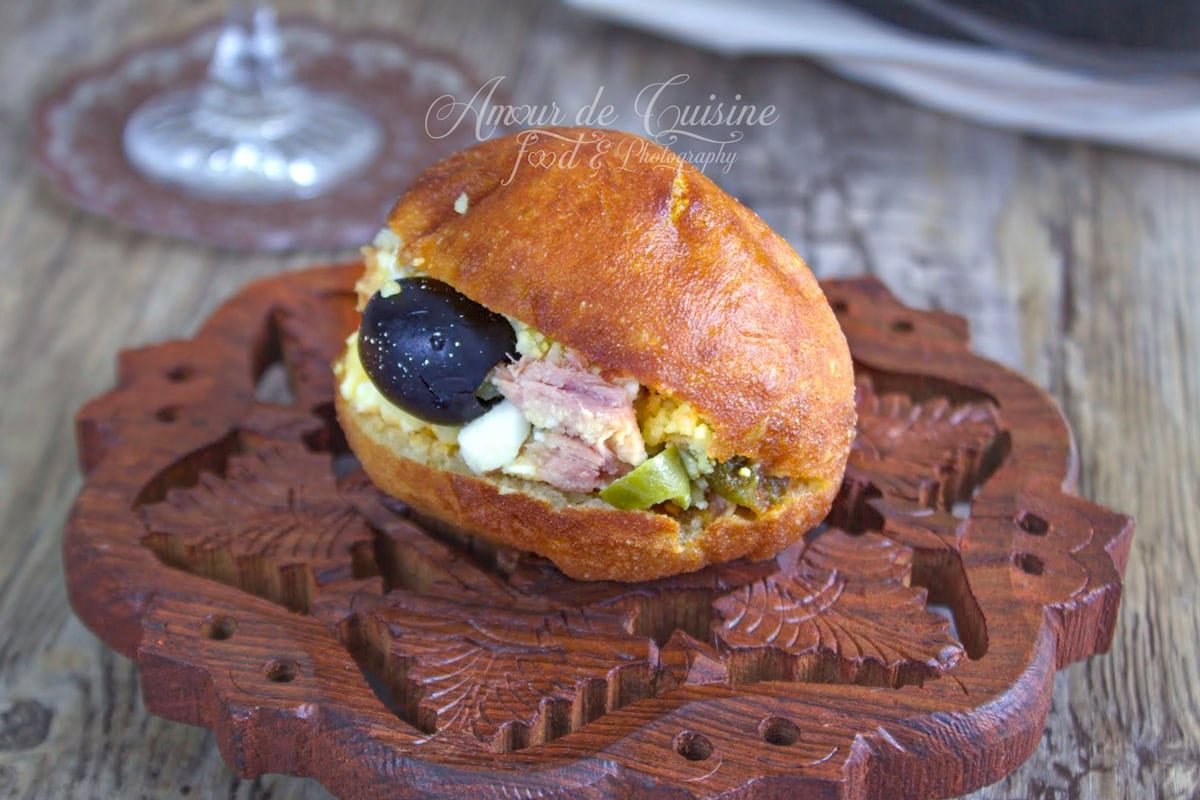Fricassé tunisien maison bien doré, garni de thon, œuf et pomme de terre, posé sur une planche en bois sculptée, avec une olive noire visible.