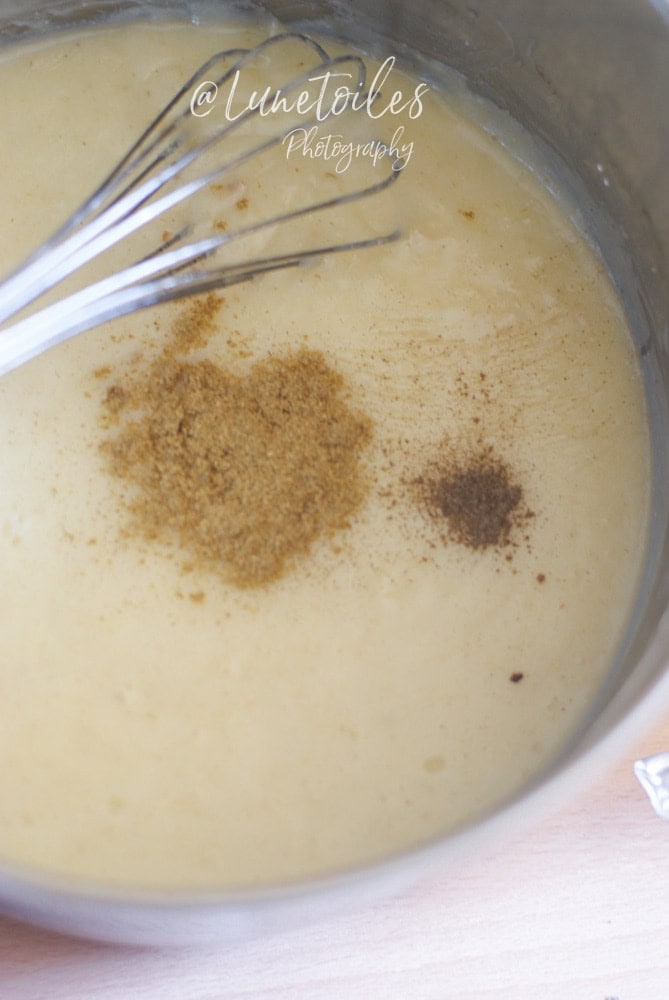 Close-up of a saucepan filled with creamy béchamel sauce being whisked, topped with freshly added nutmeg and black pepper.