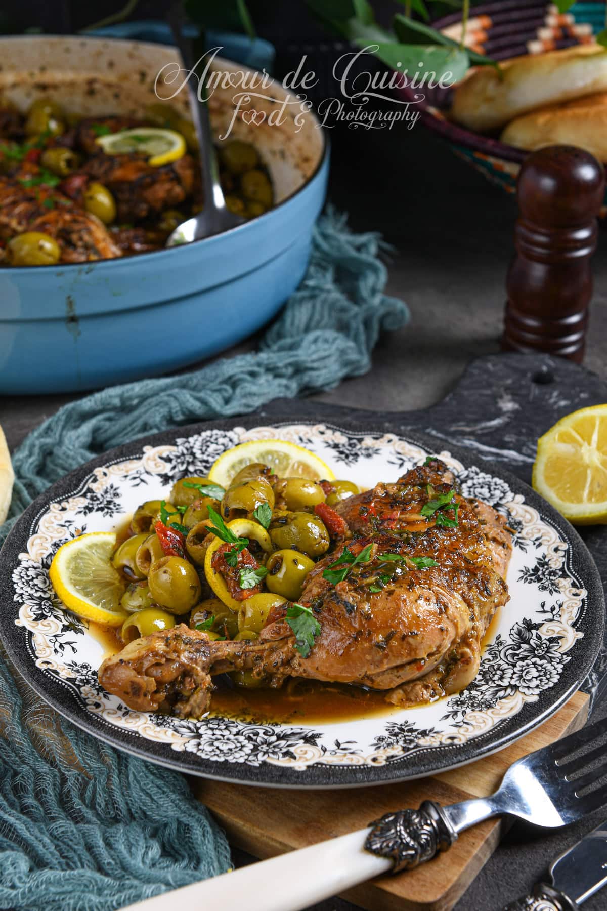 Assiette de tajine de poulet aux olives vertes, tomates séchées confites et rondelles de citron, servie avec sauce parfumée aux herbes, présentée sur une assiette décorée, avec cocotte en arrière-plan.