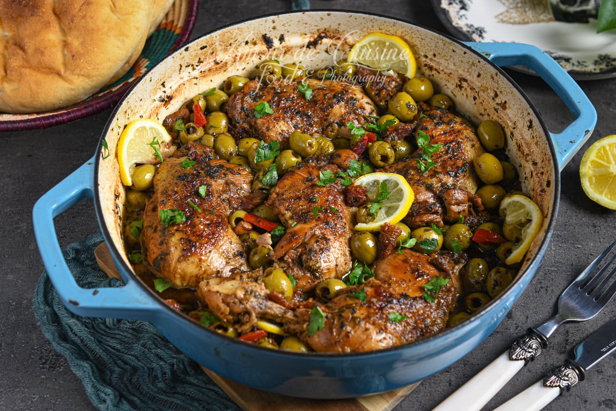 Cocotte en fonte bleue remplie de tajine de poulet aux olives vertes, tomates séchées et rondelles de citron, garnie de coriandre fraîche, servie avec pain maison