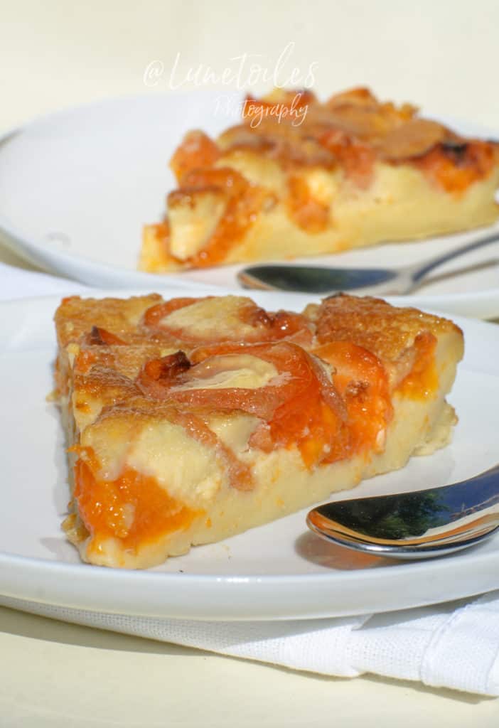 Two slices of apricot clafoutis served on white plates with teaspoons, resting on a white napkin. The clafoutis has a golden-brown top with caramelized apricot pieces embedded in a creamy, custard-like base. The dessert is well-lit, emphasizing its glossy texture and vibrant orange apricot color